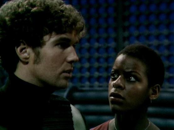 Claude 3.7 describes the image as: "This image appears to be a still from a television series, showing two individuals in what seems to be a dimly lit, futuristic setting. The scene captures an intense moment between the characters, with a blue-tinted background that suggests a spacecraft or technical environment. One person has curly hair and is wearing dark clothing, while the other is wearing what appears to be a reddish outfit. The lighting and cinematography have that distinctive vintage s…