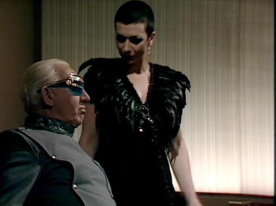 Claude 3.7 describes the image as: "This image appears to be a still from a science fiction television series from the late 1970s or early 1980s. The scene shows two people in a dramatic interaction. On the left is an older person with white hair wearing distinctive blue-tinted glasses and a gray and dark outfit with a high collar. Standing nearby is a person in a striking black feathered or textured costume with a v-neck design.

The setting appears to be an interior room with light-colored cu…