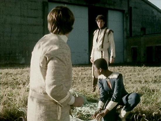 Claude Sonnet 4.0 describes the image as: "I can see a scene set in what appears to be a rural or desolate outdoor location with some kind of industrial or institutional building in the background. There are three people in the scene - one person wearing light-colored clothing in the foreground with their back to the camera, another person in similar light clothing standing in the background near the building, and a third person crouched or kneeling on the ground wearing darker clothing. The se…