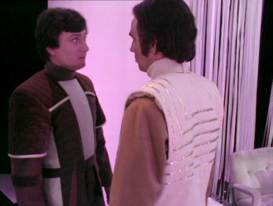 Claude 3.7 describes the image as: "The image shows two people in conversation, bathed in a purple-tinted light. They are wearing distinctive futuristic outfits typical of science fiction television productions from the late 1970s or early 1980s. The person on the left wears a brown and white color-blocked uniform, while the person on the right has on a light-colored garment with decorative striped elements. 

The setting appears to be an interior space with curtains or vertical panels visible …