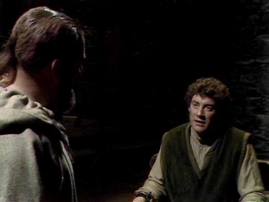 Claude 3.7 describes the image as: "The image appears to be from a television drama set in what looks like a dimly lit, rustic interior with stone walls. Two people are shown in conversation - one with curly hair wearing a green vest over light-colored clothing is positioned on the right side of the frame, while another person is partially visible from behind on the left side, wearing what appears to be a hooded or cloaked garment.

The scene has a distinctly vintage quality to its production s…