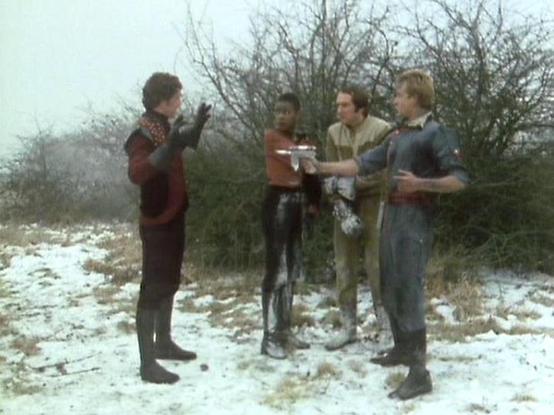 Claude 3.7 describes the image as: "This image appears to be from a science fiction television series, captured in a winter setting with snow on the ground. Four people are standing in conversation, wearing distinctive futuristic costumes that suggest a space or sci-fi theme. The costumes include dark leather-like materials and unique styling typical of 1970s/80s television productions. The scene takes place in a sparse outdoor location with bare trees and shrubs in the background, creating a s…