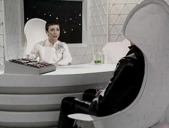Claude 3.7 describes the image as: "This image shows a futuristic, minimalist white interior setting with geometric patterns on the walls. A person in a white outfit with a corsage or decorative element sits behind a curved desk or console with what appears to be a control panel. They appear to be engaged in conversation with another figure seated in a pod-like chair, dressed in black. The stark contrast between the white environment and the black-clad figure creates a dramatic visual dynamic. …