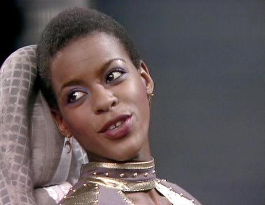 Claude 3.7 describes the image as: "The image shows a person with short dark hair and elegant earrings, looking to the side with an expressive gaze. They're wearing what appears to be a distinctive gold-colored high collar or neck piece with decorative elements, possibly part of a futuristic or sci-fi costume. The styling has a distinct retro sci-fi aesthetic characteristic of British television productions from the late 1970s or early 1980s.

The background is muted and dark, creating contrast…
