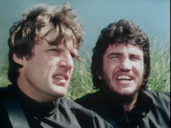 Claude 3.7 describes the image as: "The image shows two people in what appears to be an outdoor setting with tall grass or reeds in the background. The scene has a vintage film quality with a somewhat muted color palette typical of television productions from the late 1970s or early 1980s. 

Both individuals are wearing dark clothing and appear to be in the midst of an intense moment, with expressions suggesting distress or exertion. The person on the right has darker, slightly longer hair and …