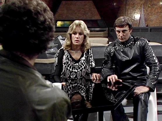 Claude 3.7 describes the image as: "This image appears to be from a vintage science fiction TV series, showing a scene in what looks like a spacecraft or futuristic control room. The setting features technical equipment and utilitarian design elements typical of sci-fi productions from the late 1970s or early 1980s.

The scene captures three individuals in conversation. On the right is a person wearing a distinctive black leather outfit with decorative studs or buttons along the sleeves. In the…