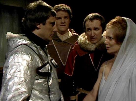 Claude 3.7 describes the image as: "The image shows a scene from what appears to be a science fiction television production from the late 1970s or early 1980s. Four people are gathered in what looks like a darkened set. On the left is a person wearing a distinctive silver/metallic costume with black trim. The others are dressed in period or theatrical costumes - one in a reddish outfit with decorative elements, another in dark clothing with fur trim, and on the right, a person wearing a white v…