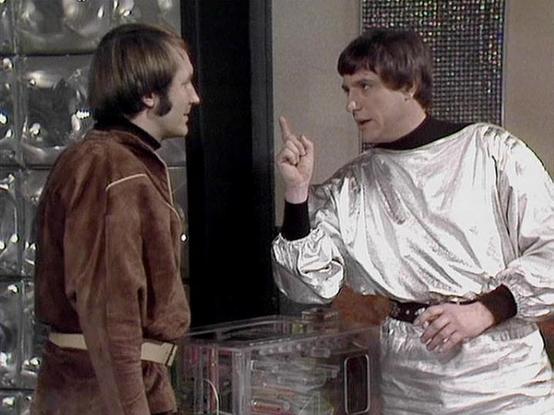 Claude 3.7 describes the image as: "The image shows a scene from a science fiction television series, featuring two people in a futuristic setting. On the right is someone wearing a distinctive silver/white outfit with a brown collar, who appears to be making a point with a raised finger during a conversation. The person on the left is dressed in a brown uniform with lighter trim. They appear to be having a discussion beside what looks like a clear console or control panel. The background featu…
