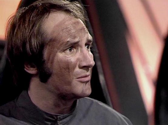 Claude 3.7 describes the image as: "This image appears to be from a science fiction television series, showing a profile view of a person in what looks like a futuristic setting. The individual is wearing what appears to be a dark, possibly uniform-like outfit. They have distinctive sideburns and a hairstyle that suggests the production is from the late 1970s or early 1980s era.

The background has a reddish-pink tint with some structural elements visible, which fits with the aesthetic of sci-f…