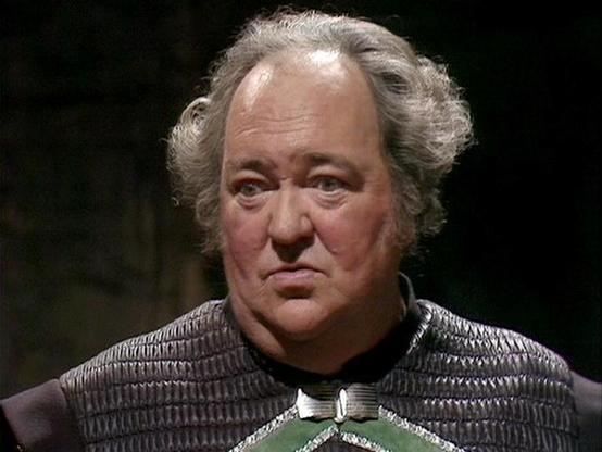 Claude 3.7 describes the image as: "The image shows a person with gray hair wearing what appears to be an elaborate costume with a textured, padded or quilted dark upper garment that has a distinctive design with a green section at the chest area. The costume has a medieval or science fiction styling to it, which is consistent with the aesthetic of classic British television drama. 

The setting appears dark, creating a dramatic atmosphere typical of studio-based productions from that era. The …