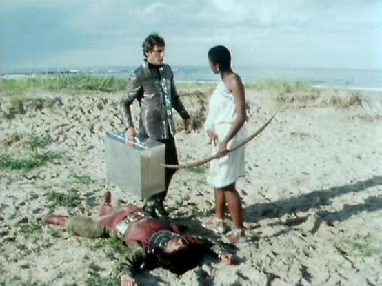 Claude 3.7 describes the image as: "This image appears to be from a television scene set on a sandy beach area. Two individuals are standing and conversing - one wearing what looks like a dark outfit and the other in a white garment holding what appears to be a curved weapon or tool. There's someone lying on the ground in reddish clothing, possibly part of a dramatic scene.

The setting shows a coastal landscape with sand dunes, some vegetation, and the ocean visible in the background under a b…