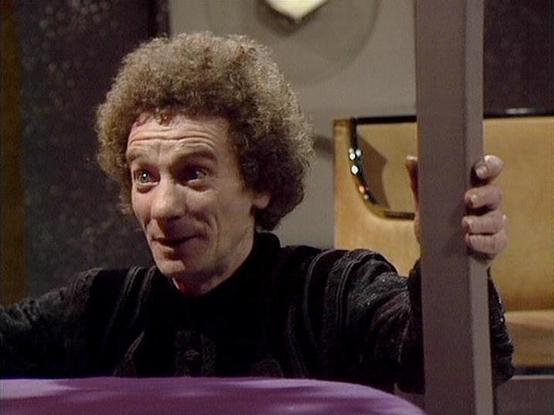 Claude 3.7 describes the image as: "This image appears to be from a classic science fiction television series from the late 1970s/early 1980s. The shot shows a person with curly hair making an expressive face while appearing to peer through or hold onto what looks like part of a set piece or doorway. They're wearing dark clothing and the image has the distinctive visual quality of productions from that era, with typical studio lighting and set design visible in the background.

The scene seems …
