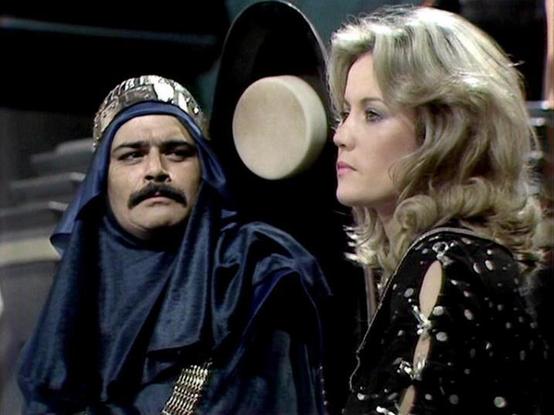 Claude 3.7 describes the image as: "The image appears to be from a science fiction television series, showing two people in a scene together. On the left is a person wearing an elaborate blue costume with a headdress that has metallic embellishments, reminiscent of a Middle Eastern or exotic styling, complete with a mustache. On the right is a person with blonde wavy hair wearing a black outfit with what looks like polka dots or small decorative elements.

The setting seems to be some kind of f…