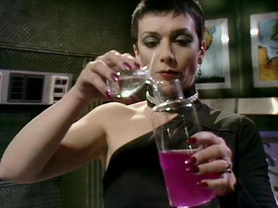Claude 3.7 describes the image as: "This image appears to be from a science fiction television series, showing a scene in what looks like a futuristic laboratory or spaceship interior. The person in the image has short dark hair and is wearing a dark outfit with a distinctive collar. They are carefully pouring a bright pink or magenta liquid from a small glass container into a larger beaker or flask. The vibrant color of the liquid suggests it might be some kind of special chemical, potion, or …
