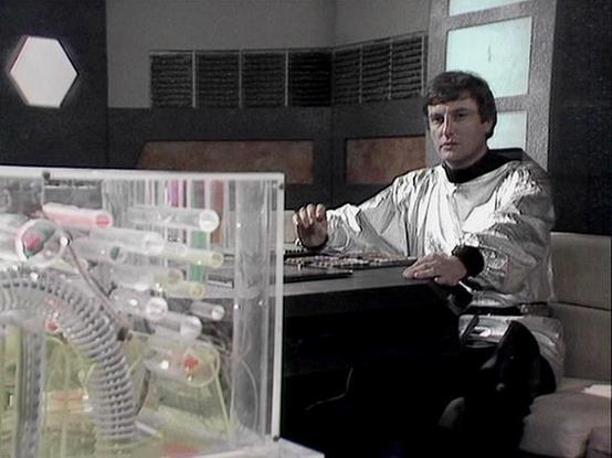 Claude 3.7 describes the image as: "This image shows a futuristic sci-fi control room or laboratory setting. A person is seated at what appears to be a control station, wearing a distinctive silver/white jacket with dark pants. The metallic, reflective fabric of the outfit suggests a futuristic or space-age aesthetic typical of science fiction productions from the late 1970s or early 1980s.

In the foreground, there's a transparent console or equipment case containing various tubes, wires, and …