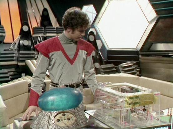 Claude Sonnet 4.0 describes the image as: "I can see this is a scene set in what appears to be a futuristic spaceship interior, with characteristic sci-fi design elements including hexagonal wall panels and advanced-looking control surfaces. The setting features sleek, technological architecture typical of space-based science fiction productions from this era.

The scene shows someone in a distinctive costume - a grey and red outfit that appears to be some kind of uniform or specialized clothin…