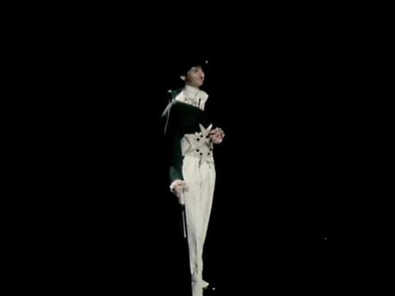 Claude Sonnet 4.0 describes the image as: "I can see this is a dramatic scene set against a stark black background, creating a theatrical, minimalist atmosphere. The figure is wearing an elegant period costume consisting of a dark jacket or coat paired with light-colored trousers, suggesting a formal or aristocratic character. The costume design has a historical feel, possibly 17th or 18th century inspired, with what appears to be decorative elements on the clothing. The dramatic lighting and s…