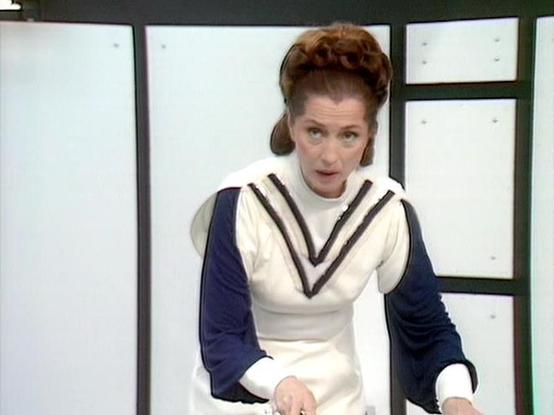 Claude 3.7 describes the image as: "This image shows a person with an upswept hairstyle wearing what appears to be a distinctive costume with a white tunic featuring a navy blue and black V-shaped collar design. The outfit has navy blue sleeves contrasting with the white body of the garment. They appear to be in what looks like a futuristic or stylized set with white panels and dark green or black framing visible in the background. The aesthetic suggests this is from a science fiction televisio…