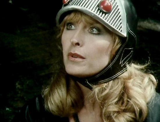 Claude Sonnet 4.0 describes the image as: "I can see this is a scene featuring a character wearing a distinctive futuristic helmet with striped patterns and what appears to be some kind of technological or decorative elements. The character has blonde, wavy hair and is wearing what looks like a fur-collared garment. The lighting and cinematography are characteristic of late 1970s British science fiction television, with dramatic lighting against a dark background. The helmet design suggests thi…