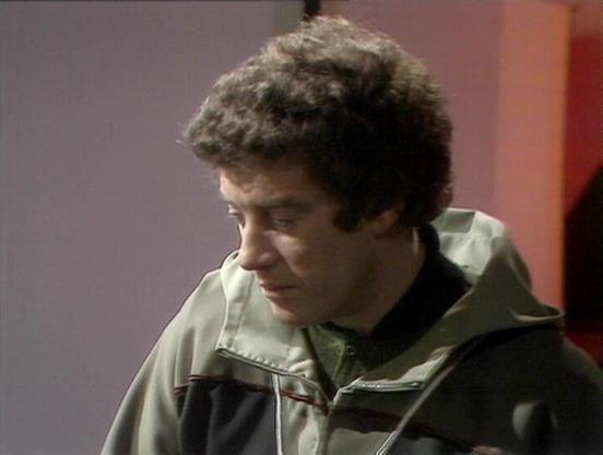 Claude Sonnet 4.0 describes the image as: "I can see this is a scene from a science fiction television series, showing someone wearing what appears to be a light-colored, military or utilitarian-style jacket or uniform. The setting has the distinctive look of late 1970s/early 1980s television production, with characteristic lighting and background colors typical of that era's sci-fi shows. The person appears to be in some kind of indoor setting, possibly aboard a spacecraft or in a futuristic f…