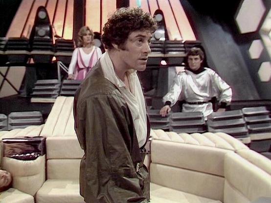 Claude 3.7 describes the image as: "This image shows a scene from a science fiction television series filmed in the late 1970s or early 1980s. The setting appears to be the interior of a spacecraft or space station with a distinctive futuristic design featuring hexagonal wall patterns and tiered levels.

In the foreground, there's a person wearing a brown jacket over light-colored clothing, standing near what looks like cream-colored seating. In the background, two other people are visible on e…