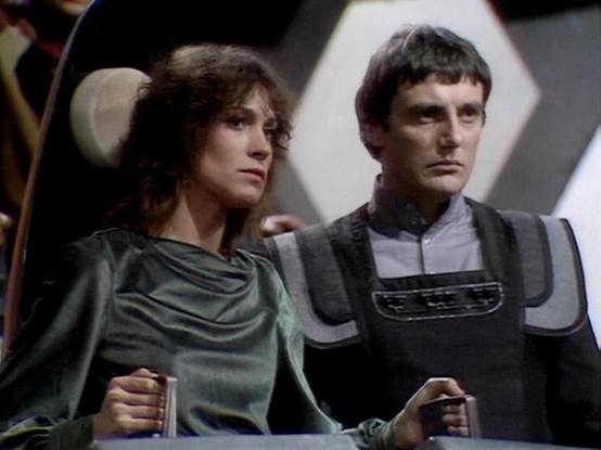 Claude Sonnet 4.0 describes the image as: "This appears to be a scene from a science fiction television series, showing two characters in what looks like the interior of a spacecraft or futuristic facility. The setting has a distinctly retro-futuristic aesthetic typical of British sci-fi productions from the late 1970s or early 1980s.

The scene shows two people seated together - one wearing a flowing green/gray draped garment, and another in what appears to be a dark vest or tunic-style outfit…