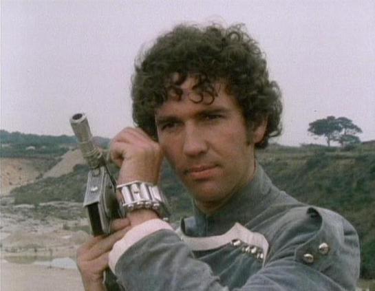 Claude 3.7 describes the image as: "The image shows a person with curly dark hair in what appears to be an outdoor, rugged landscape setting. They're wearing what looks like a gray uniform or jacket with metallic accents, typical of science fiction costume design from the late 1970s or early 1980s. The individual is holding what appears to be a prop weapon, possibly a futuristic gun or blaster.

The scene has a distinctly retro sci-fi aesthetic with a barren, possibly quarry-like landscape in t…