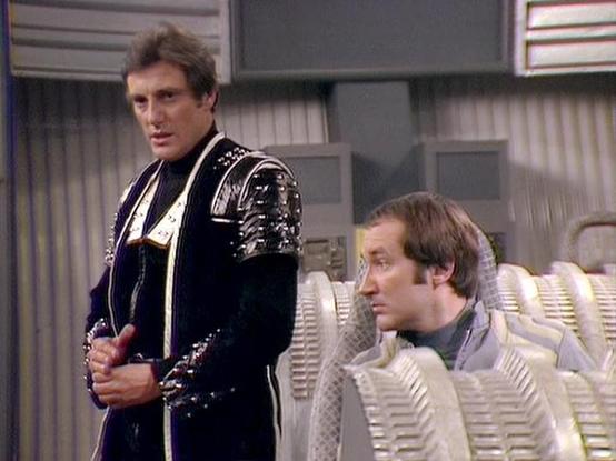 Claude Sonnet 4.0 describes the image as: "This scene shows two characters in what appears to be the interior of a spacecraft or space station. The setting features a futuristic, sterile environment with white and metallic surfaces typical of science fiction television production design from the late 1970s or early 1980s.

One character is standing and wearing an elaborate black costume with ornate detailing, silver or metallic embellishments, and what appears to be a formal, almost ceremonial …