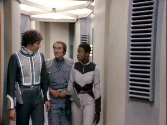Claude Sonnet 4.0 describes the image as: "This scene shows three crew members in what appears to be a corridor or passageway aboard their spacecraft. The setting features the series' characteristic clean, futuristic interior design with white walls and geometric lighting panels in the ceiling. The characters are wearing the show's distinctive costume designs - form-fitting outfits in different color schemes that were typical of late 1970s science fiction television. One person wears a teal and…