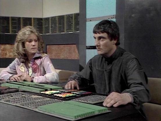 Claude Sonnet 4.0 describes the image as: "This scene shows two characters in what appears to be the control room or bridge of a spacecraft. The setting features a futuristic control console with various buttons, switches, and displays typical of science fiction television from the late 1970s. The console has a distinctive green section and multiple control panels with colorful indicators.

The woman is wearing a light pink or cream-colored outfit and has blonde, feathered hair characteristic o…