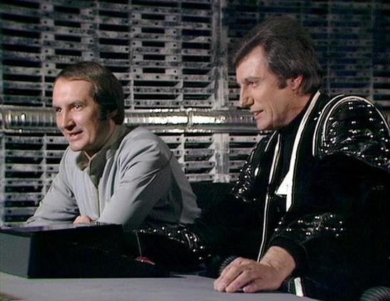 Claude 3.7 describes the image as: "The image shows a scene from a classic science fiction television series from the late 1970s/early 1980s. It depicts two characters in what appears to be a futuristic control room or spacecraft setting, with technical equipment and computer banks visible in the background.

On the left is a person wearing a light gray outfit, while on the right is someone dressed in a distinctive black leather or vinyl costume with silver accents. Both appear to be seated at …