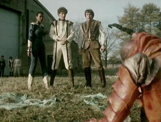 Claude 3.7 describes the image as: "This image appears to be from a science fiction television series, showing three people standing in a field near a building. They're wearing distinctive futuristic costumes - one in a dark outfit with light-colored boots, and two others in light-colored tunics with embellishments and boots, suggesting a sci-fi or fantasy setting.

In the foreground, there's a partial view of what looks like someone's hand or arm in a reddish/brown leather glove or sleeve. The…