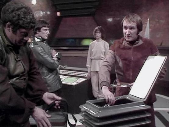 Claude Sonnet 4.0 describes the image as: "This appears to be a scene set aboard a spacecraft, likely in a control room or technical area. The setting has the characteristic futuristic design elements typical of 1970s science fiction television, with curved walls and control panels. Four characters are present in the scene, gathered around what appears to be some kind of technical equipment or container on a table. The lighting and overall aesthetic suggests this is from a dramatic moment in th…