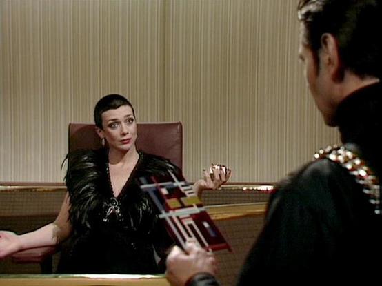 Claude 3.7 describes the image as: "The image shows a scene from what appears to be a science fiction television series from the late 1970s or early 1980s. It depicts a formal meeting or confrontation in what looks like a governmental or administrative setting. 

In the foreground, a person with short dark hair wearing an elegant black outfit with feathered or textured shoulders sits in a leather chair. They appear to be in a position of authority, gesturing while speaking. The costume has a dr…