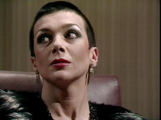 Claude 3.7 describes the image as: "This image appears to be from a television series, showing a person with a very short, dark haircut and dramatic makeup including defined eyebrows and red lipstick. They are wearing elegant dangling earrings and what looks like a dark garment with a textured or fur-like collar. The person is looking upward, creating a striking and somewhat imperious expression.

The setting appears to be an interior scene with neutral-colored walls, and there's what looks lik…