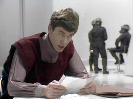 Claude Sonnet 4.0 describes the image as: "I can see this appears to be from a science fiction television series, showing a scene in what looks like a sterile, white institutional setting. In the foreground, there's a person in dark clothing reading or examining some papers at what appears to be a desk or table. In the background, there are figures in what look like military or security uniforms. The setting has a clinical, authoritarian atmosphere typical of dystopian sci-fi productions from t…