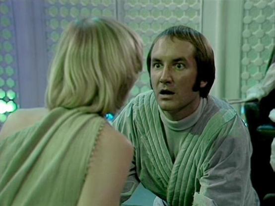 Claude Sonnet 4.0 describes the image as: "I can see this appears to be from a science fiction television series, showing two people in what looks like a futuristic interior setting with distinctive hexagonal or geometric patterns visible in the background. One person has blonde hair and is wearing what appears to be a light-colored garment, while the other person has dark hair and is wearing what looks like a ribbed or textured outfit in muted tones. The setting has a clinical or technological…