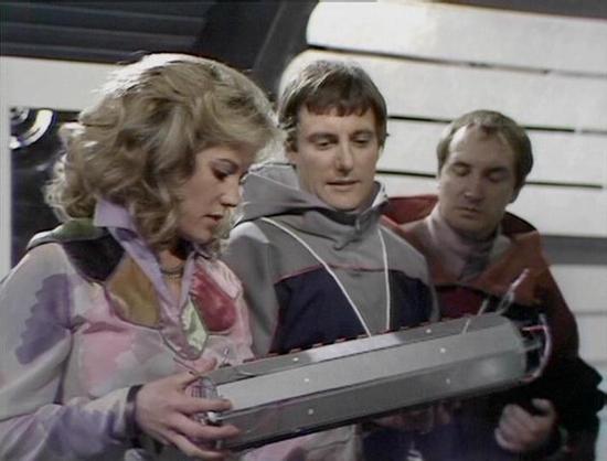 Claude 3.7 describes the image as: "This image appears to be from a classic science fiction television series from the late 1970s or early 1980s. It shows three people in a futuristic spacecraft setting, wearing distinctive space-age costumes with metallic and colored accents typical of the era's sci-fi aesthetic.

In the foreground, a woman with blonde curly hair is examining what appears to be some kind of electronic device or control panel. She's wearing a pinkish-lavender outfit. Two men in…