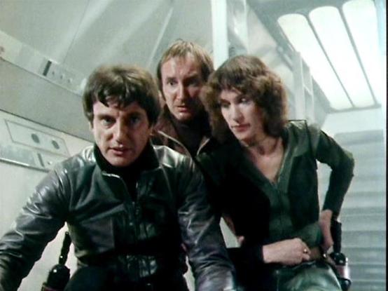 Claude 3.7 describes the image as: "The image shows three people in a sci-fi corridor setting with bright, minimalist futuristic design featuring white walls and overhead lighting. They're wearing distinctive leather-like costumes typical of late 1970s/early 1980s British science fiction television. The person on the left wears a dark leather jacket, the middle figure is partially visible between the others, and the person on the right wears a green outfit. They appear to be in some kind of spa…