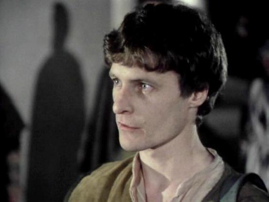 Claude 3.7 describes the image as: "This image appears to be from a film or television production with a period or science fiction setting. The image shows a person with dark, tousled hair in profile, looking thoughtfully to the side. They're wearing what looks like a simple beige or light-colored shirt with what might be a dark vest or suspender. 

The lighting creates a dramatic atmosphere with shadows visible on the wall behind, suggesting this might be from a dimly lit scene. The muted colo…