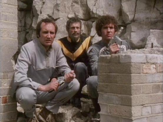Claude 3.7 describes the image as: "The image shows three people crouched among ruins or debris of what appears to be a damaged building with stone walls. They're wearing futuristic-looking costumes typical of science fiction television from the late 1970s/early 1980s era. The person on the left wears a gray outfit with darker gray accents, the middle figure has on a black and yellow/gold top, and the right figure is dressed in gray. They seem to be hiding or taking cover behind some brick stru…