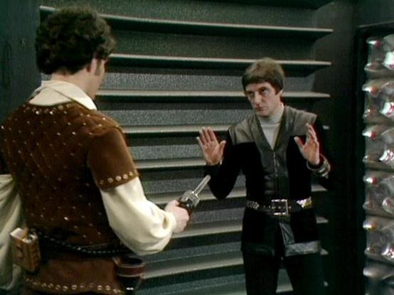 Claude Sonnet 4.0 describes the image as: "This scene appears to be set in a futuristic interior, likely aboard a spacecraft, with characteristic metallic wall panels and angular architecture typical of science fiction productions from this era. Two men are engaged in what appears to be a tense confrontation. One figure is dressed in period-style clothing with ornate brown leather studded with metal details over a white shirt, while the other wears dark, fitted clothing with a high collar. The …