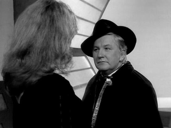 Claude Sonnet 4.0 describes the image as: "I can see this is a black and white image showing two people in what appears to be a dramatic scene. One person is shown from behind with shoulder-length hair, while the other is wearing a dark hat and coat, giving them a distinctive period appearance. The setting appears to be indoors with some architectural elements visible in the background. The lighting and composition suggest this is from a television production, with the formal costume styling in…