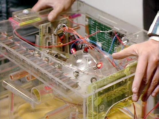 Claude 3.7 describes the image as: "The image shows a transparent electronic device or prototype being worked on by hands visible from both sides. This appears to be a clear plastic case containing circuit boards, wires in various colors (red, blue, yellow), and what looks like a central mechanical component with LEDs or small lights visible. The internal components are exposed, suggesting it might be an experimental device or prop used in a science fiction television production. The transparen…
