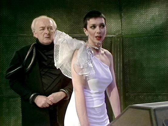Claude 3.7 describes the image as: "The image shows a scene set in what appears to be a futuristic space setting with distinctive green metallic wall panels in the background. The scene features two individuals in what seems to be a formal or dramatic moment. 

On the left is an older person wearing dark clothing, possibly a black turtleneck or jacket. On the right is someone in an elegant, light-colored satin dress or gown with a one-shoulder design and some sparkly embellishment or accessory.…