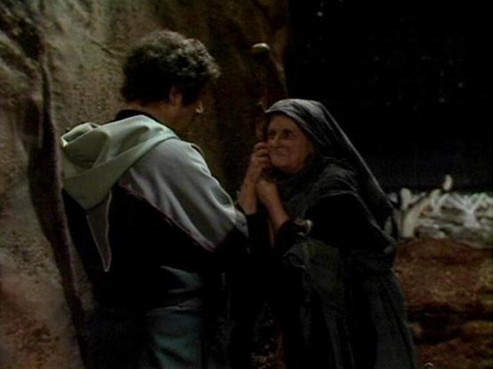 Claude Sonnet 4.0 describes the image as: "I can see this appears to be a scene set in what looks like a cave or underground location with rough stone walls. Two figures are present - one wearing lighter colored clothing and another in darker robes with a head covering. The setting has a rustic, primitive quality with what appears to be some kind of bedding or supplies visible in the background. The lighting is dim and atmospheric, creating shadows that emphasize the cave-like environment. The …