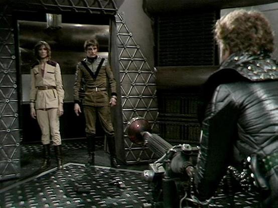 Claude 3.7 describes the image as: "This image appears to be from a classic science fiction television series, showing a scene set in what looks like a futuristic or spacecraft interior. The setting features metallic walls with a triangular lattice pattern and a distinctive industrial design aesthetic typical of 1970s/80s sci-fi productions.

Several people are visible in the frame - two standing in what appears to be a doorway or elevator (one in a beige/cream uniform and another in an olive/b…