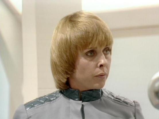 Claude Sonnet 4.0 describes the image as: "I can see this is a scene from a science fiction television series, showing a person with blonde hair wearing what appears to be a futuristic uniform or costume with grey and green coloring and distinctive shoulder details. The setting appears to be an interior space with white or light-colored walls, consistent with the sterile, spaceship-like environments typical of sci-fi productions from this era. The costume design and production values are charac…