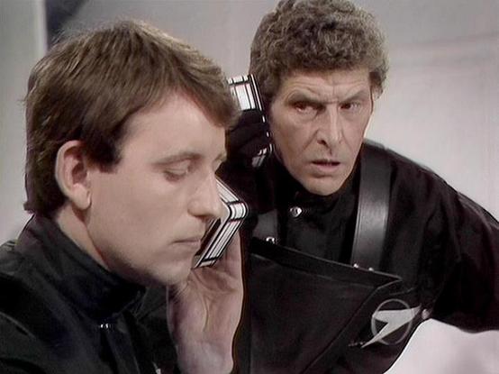 Claude Sonnet 4.0 describes the image as: "I can see this appears to be from a science fiction television production, showing two men in what looks like a tense dramatic scene. One man is wearing dark clothing and appears to be in distress or being restrained, while another figure in what appears to be black leather or similar material is positioned behind him. The setting appears to be an interior space with institutional or futuristic styling typical of sci-fi productions from this era. The l…