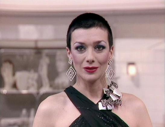 Claude 3.7 describes the image as: "The image shows a person with short dark hair wearing elegant evening attire including a black cross-body dress or top. They're accessorized with striking geometric diamond-shaped earrings and what appears to be a decorative brooch or corsage on the shoulder. Their makeup is formal with defined eyes and red lipstick.

The setting appears to be an interior space, possibly a formal venue or reception area with soft lighting visible in the background. The atmosp…